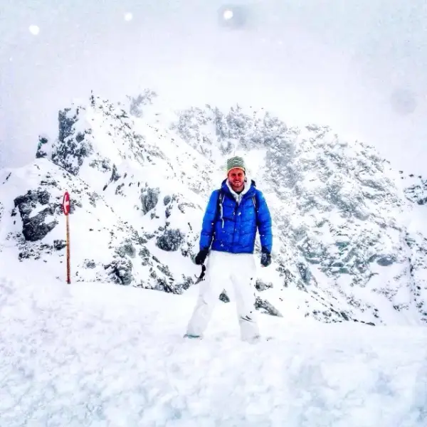 Owain in a blue down jacket standing in deep snow on a mountain in Austria