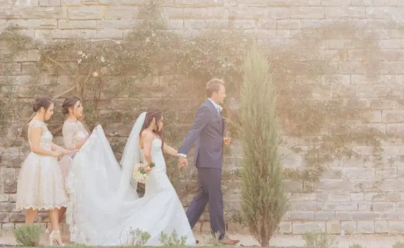 Owain walking hand in hand with his bride past a stone wall, followed by two bridesmaids carrying her train at Elmore Court